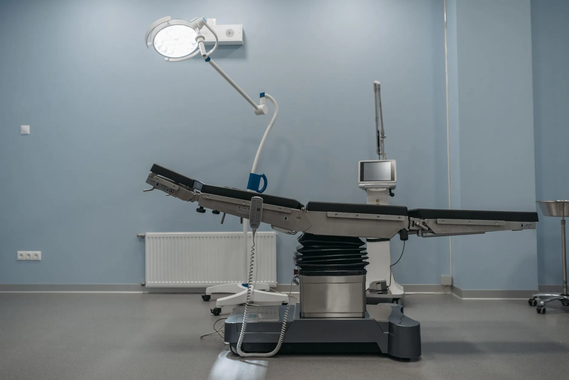 Sala de procedimentos com mesa cirúrgica, foco de luz no teto e monitor, sem pacientes