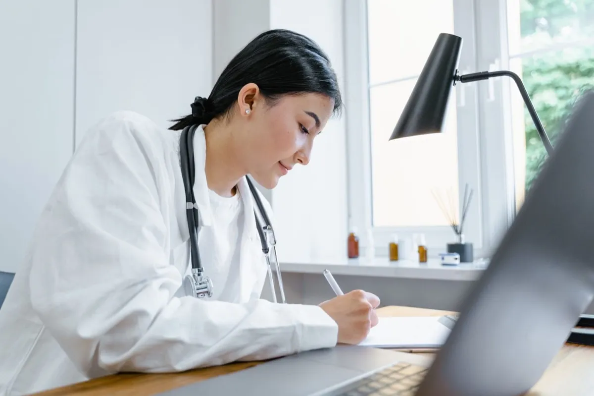 Médica de jaleco fazendo anotações à mesa, ao lado de um notebook