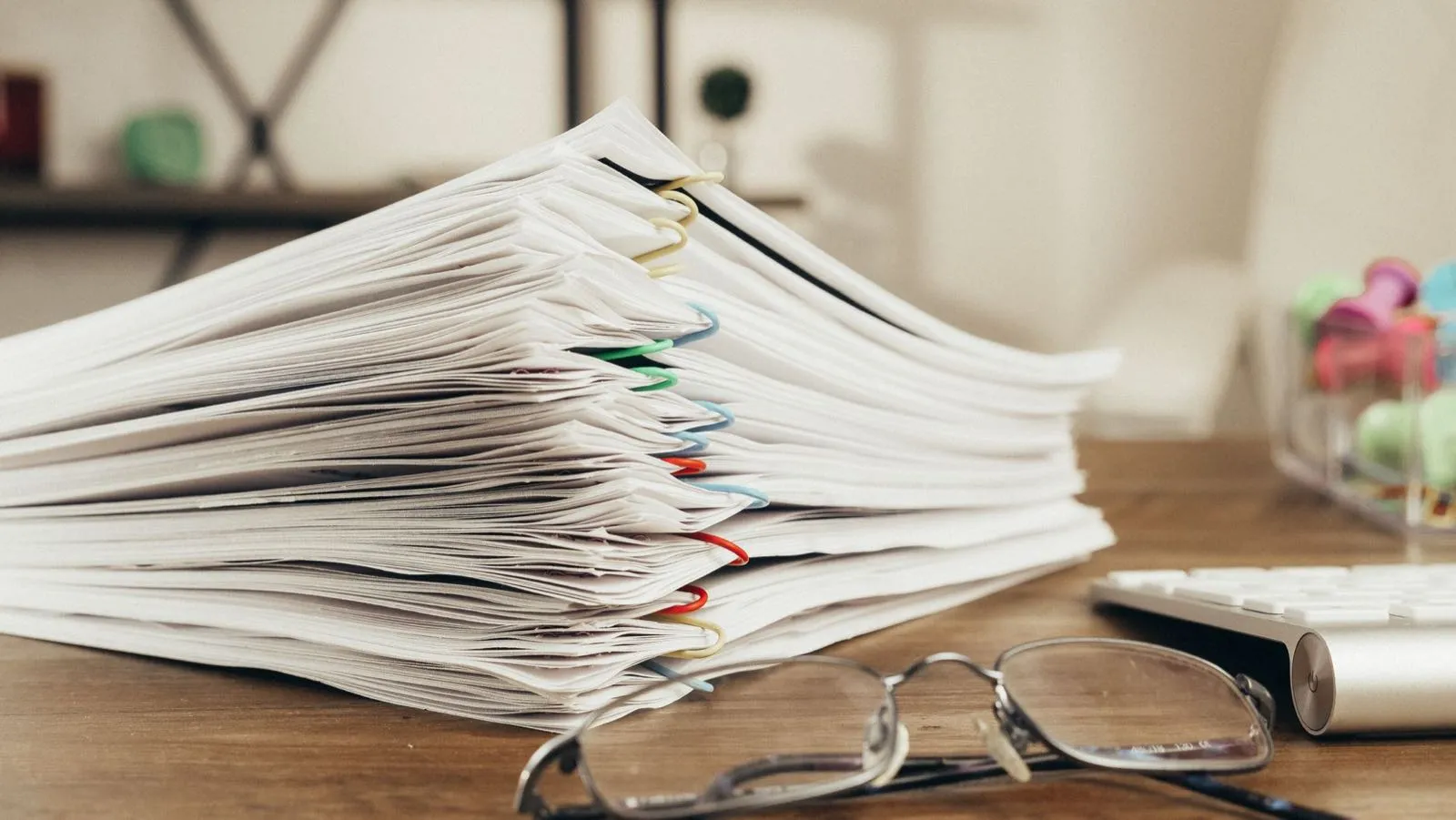 Pilha alta de papéis presos com clipes coloridos sobre uma mesa de escritório, com óculos à frente