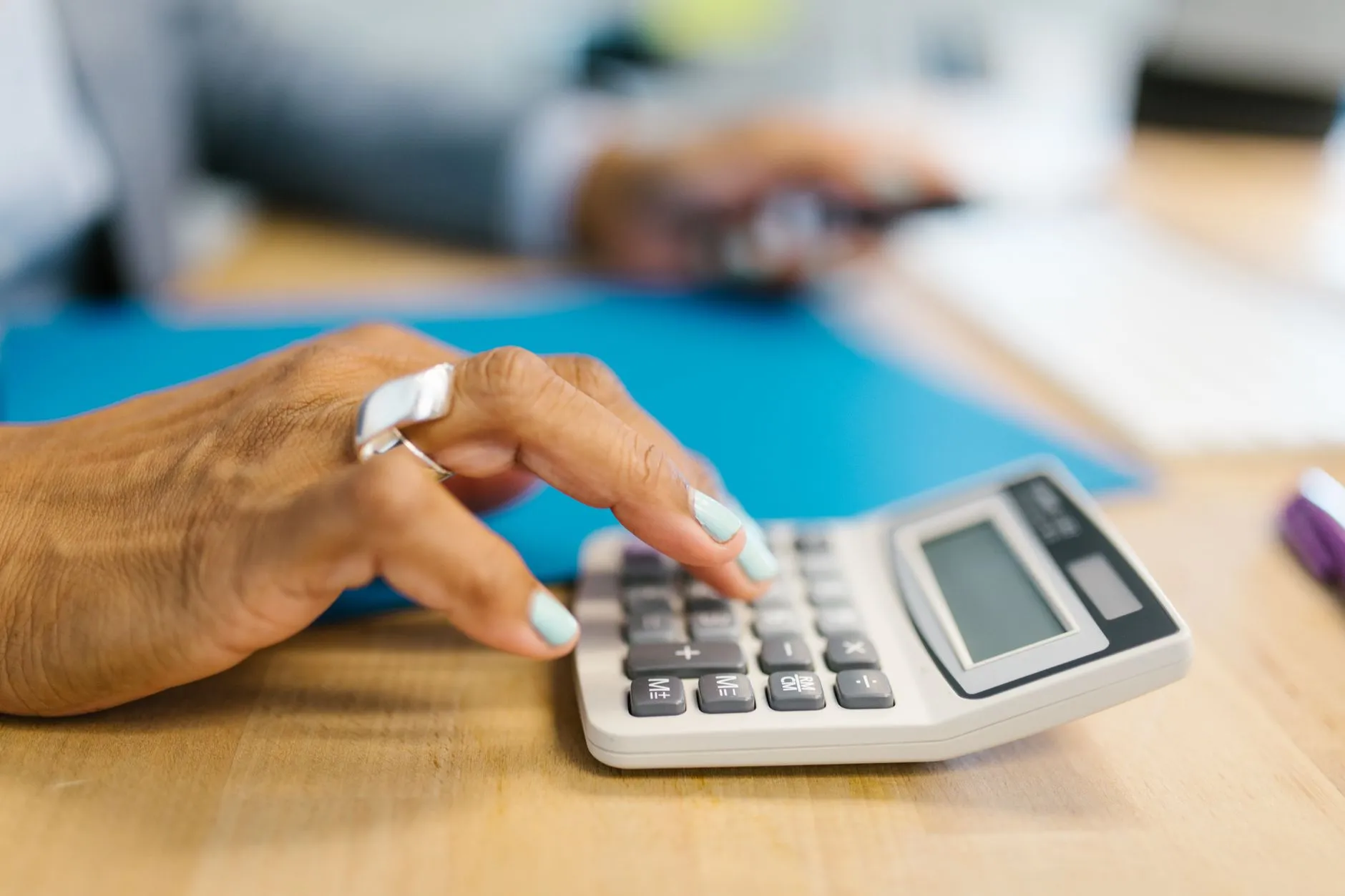 Mão com anel prateado digitando numa calculadora sobre a mesa, ao lado de uma pasta azul