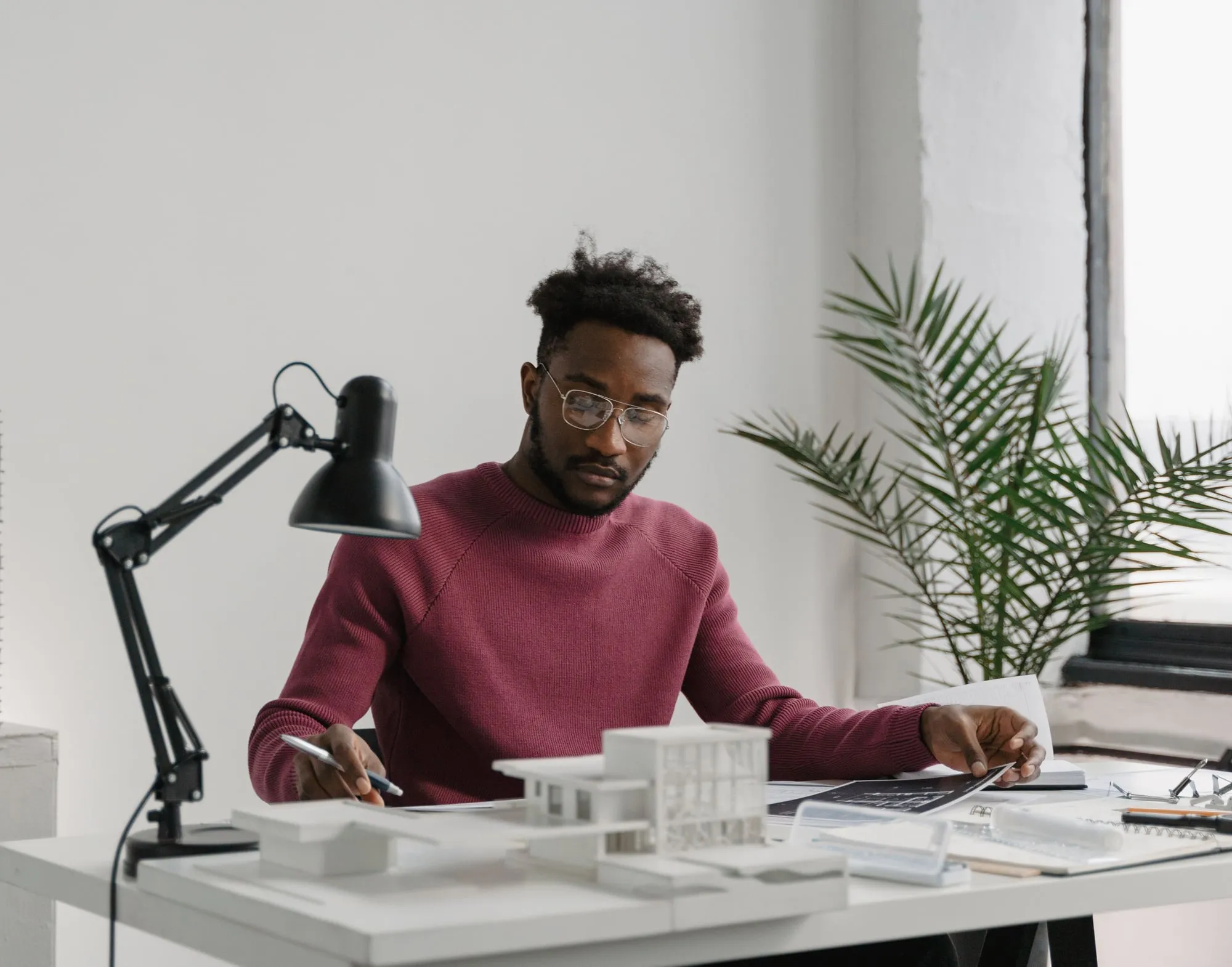 Profissional de óculos conferindo um documento à mesa do próprio escritório, com a maquete de um prédio à frente, uma luminária e uma janela ao lado